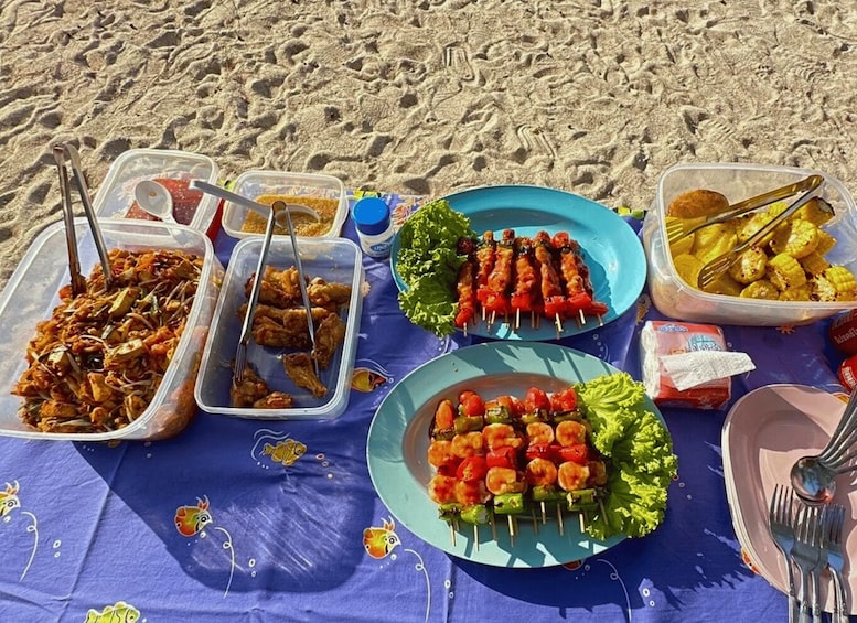 Rannalla järjestetty piknik, jossa on tarjolla monenlaista ruokaa ja ruokailuvälineet on aseteltu siniselle pöytäliinalle. 3