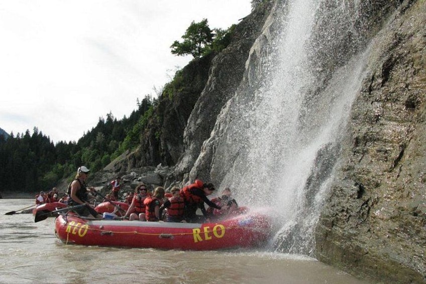 A group of people in a red raft with 'REO' on it, navigating a river near a waterfall. 5