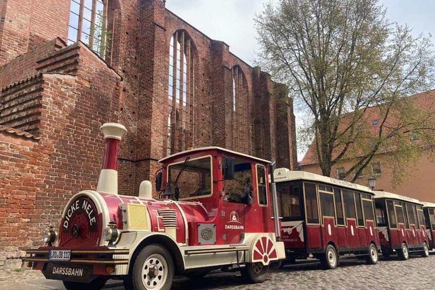 A red and white vintage train with 'DIECKE NEL' written on the front, parked in front of a brick building with arched windows. 1