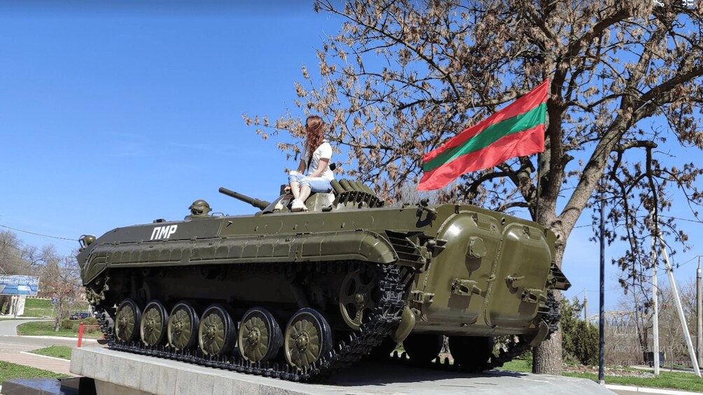 A military tank with a flag, a woman sitting on it, and a clear blue sky. 2