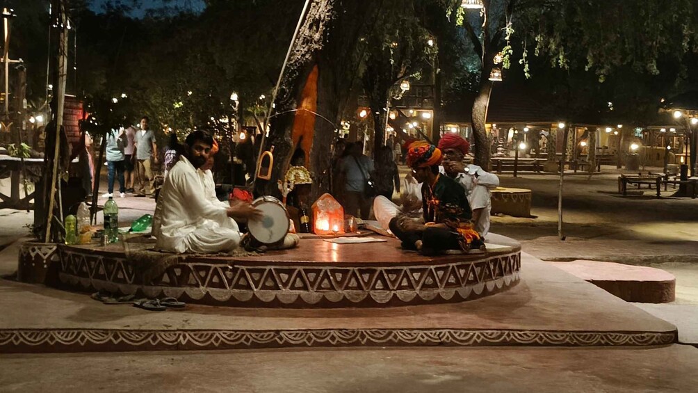 A group of people seated on a decorated platform with a fire in the center, surrounded by trees and illuminated by ambient lighting. 4