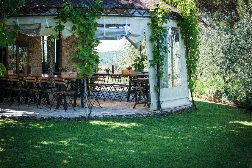 A covered outdoor dining area with wooden tables and chairs, surrounded by greenery and a stone wall. 3