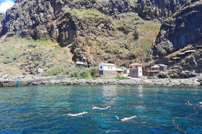 A coastal scene with swimmers in the water, rocky cliffs, and buildings on the shore. 2