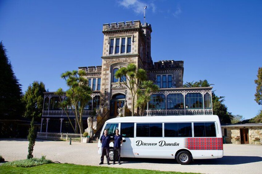 En vit buss med texten ”Upptäck Dunedin” på sidan stod parkerad framför en historisk byggnad med ett torn. 1