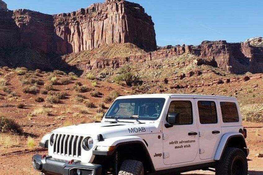 Un Jeep blanco con el texto 'adventure adventures' en la carrocería aparcado en un desierto con formaciones rocosas rojas al fondo. 5