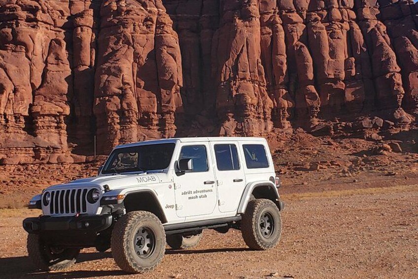 A white Jeep with 'MOAB' and 'adventure mead utah' on its side, parked in a desert with red rock formations in the background. 5