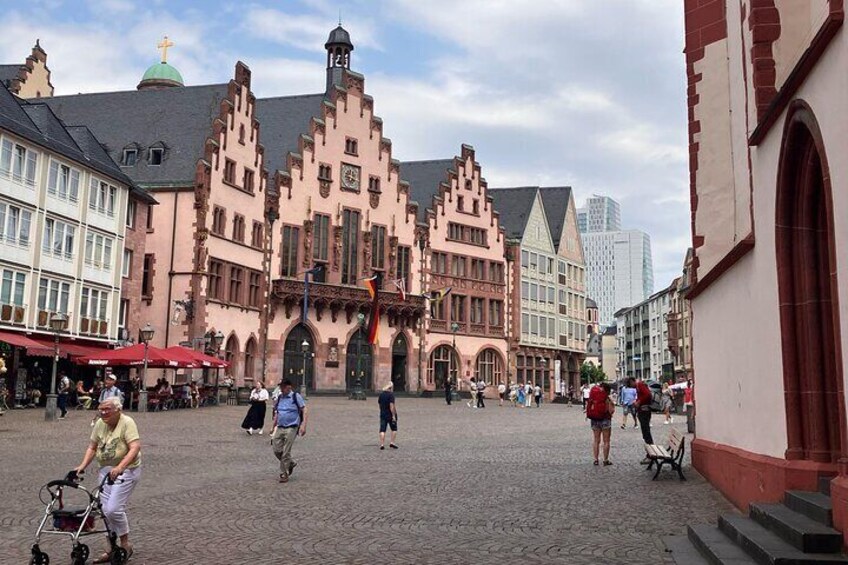 A cobblestone square with historic buildings, a clock tower, and people walking. 5