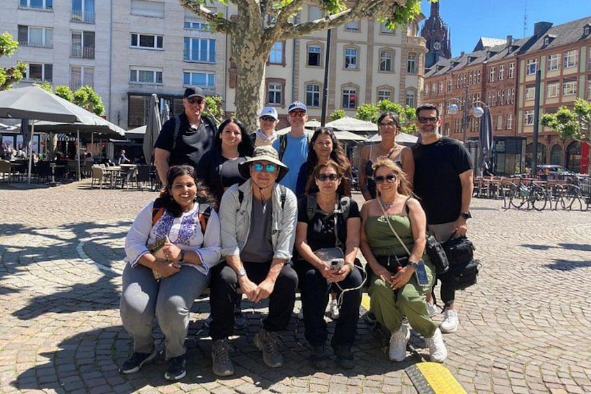 A group of people posing for a photo in a cobblestone square with buildings and trees in the background. 1
