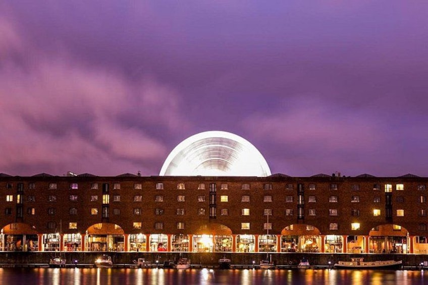 A waterfront building with illuminated arches and a large circular structure in the background. 3