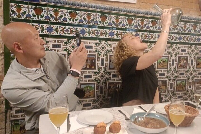 Dos personas sentadas a una mesa con comida y bebidas, una sosteniendo una cámara. 1