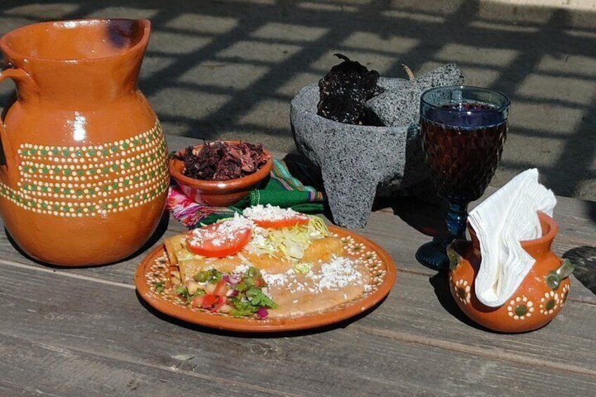 A rustic outdoor setting with a clay pot, a plate of food, a stone mortar, and a glass. 3