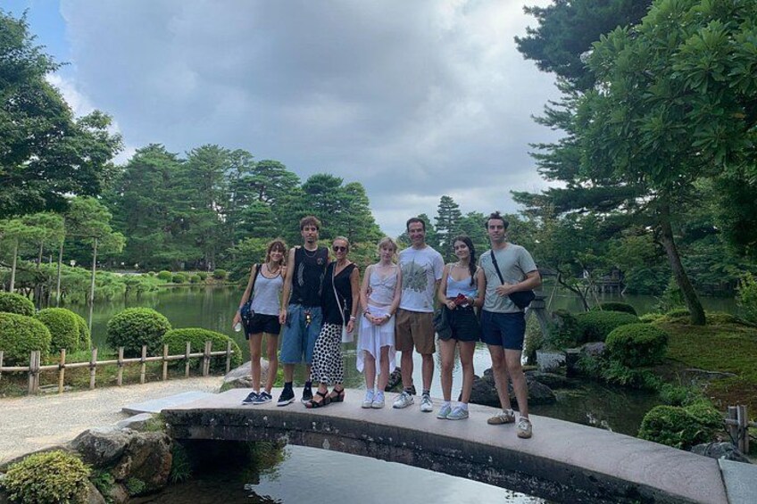 Un groupe de personnes se tient sur un pont surplombant un plan d’eau dans un parc, avec des arbres et un ciel nuageux. 2