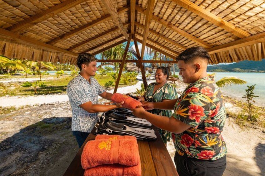 A wooden structure with a thatched roof, a wooden table with a red towel, and a beach with palm trees and water in the background. 3
