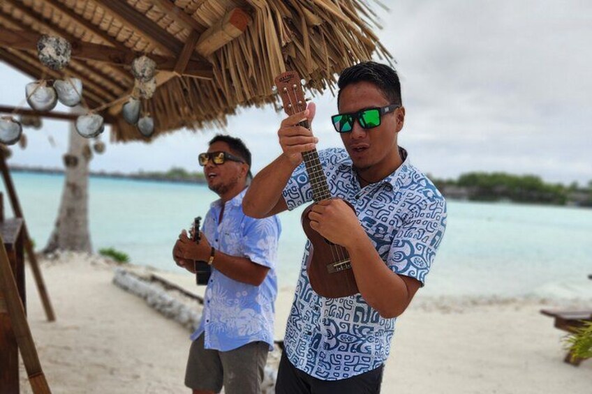Två personer spelar ukulele på en strand, och i bakgrunden syns en byggnad med ett halmtak. 2