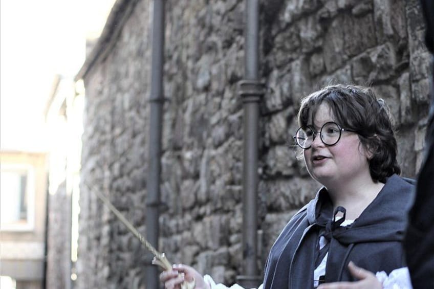 A person standing outdoors, holding a wand-like object, with a stone wall in the background. 5