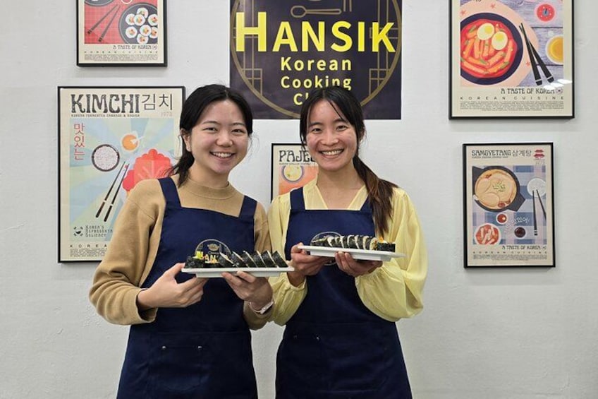 Dos personas sosteniendo platos de comida frente a una pared con carteles de cocina coreana. 2