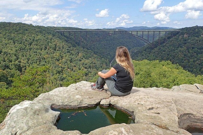 A person sitting on a rock overlooking a forested valley with a bridge in the distance. 2