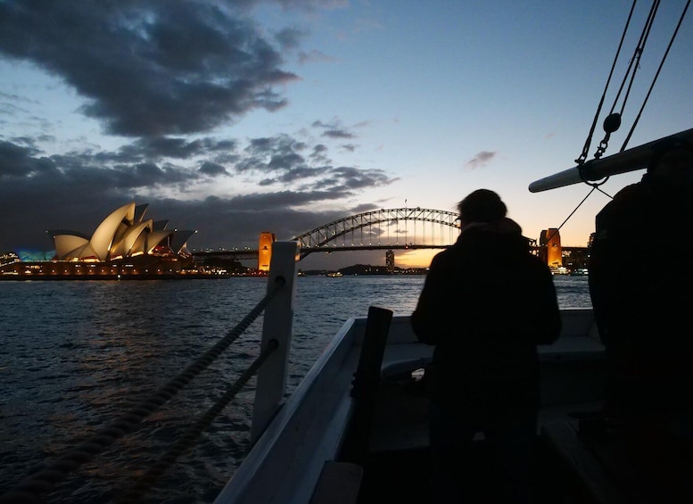 En siluett av en person på en båt, Sydneyoperahuset och Sydney Harbour Bridge i skymningen. 2
