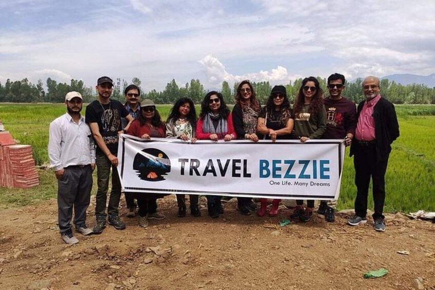 A group of people holding a banner with the text 'TRAVEL BEZZIE' and 'One Life, Many Dreams' in a rural setting with fields and a clear sky. 1