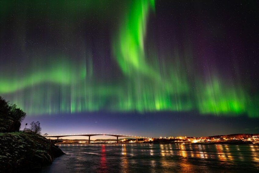 Nordlys over en flod med en bro og bylys i baggrunden. 2