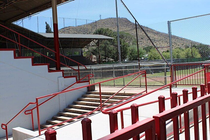 Una escalera con barandillas rojas que conduce a un campo deportivo con valla de malla y una colina al fondo. 2
