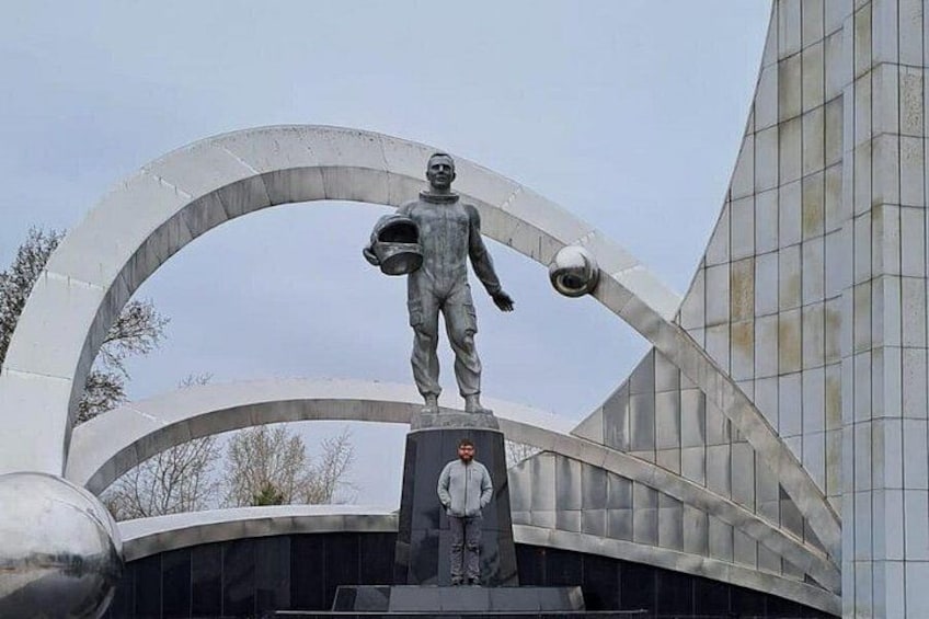 A statue of a figure holding a helmet and a trophy, standing on a pedestal with a modern building in the background. 1