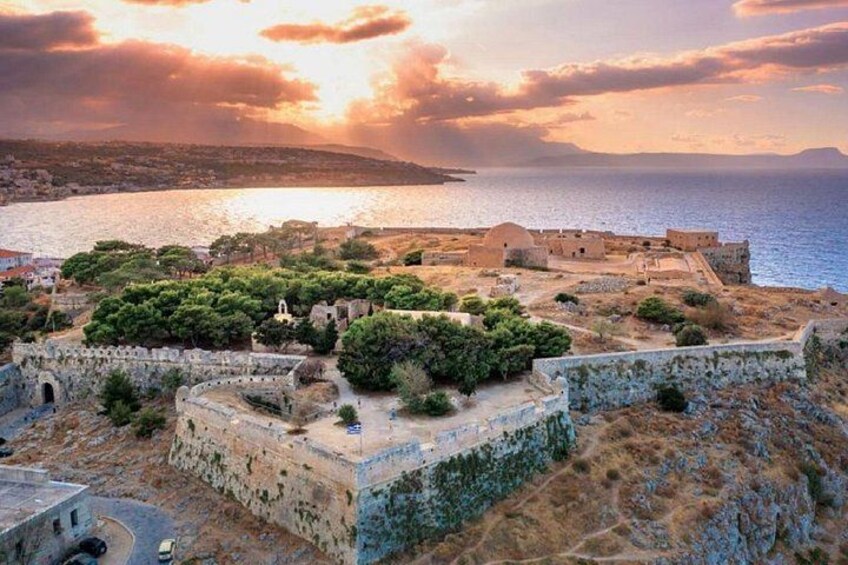 A coastal fortification with surrounding greenery and a sunset over the sea. 2