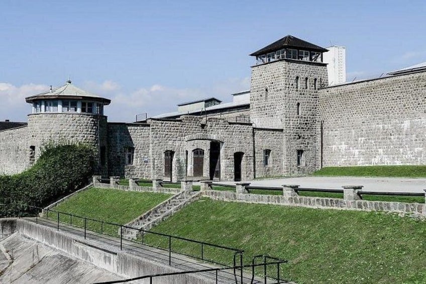 A historic stone building with a round tower and a watchtower, surrounded by a grassy area and a paved walkway. 1