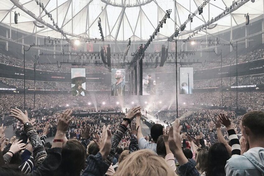 A large indoor stadium with a crowd of people, stage lighting, and a large screen displaying a performer. 4