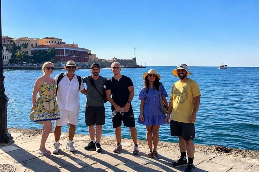 A group of people standing by a waterfront with buildings and a ship in the background. 1