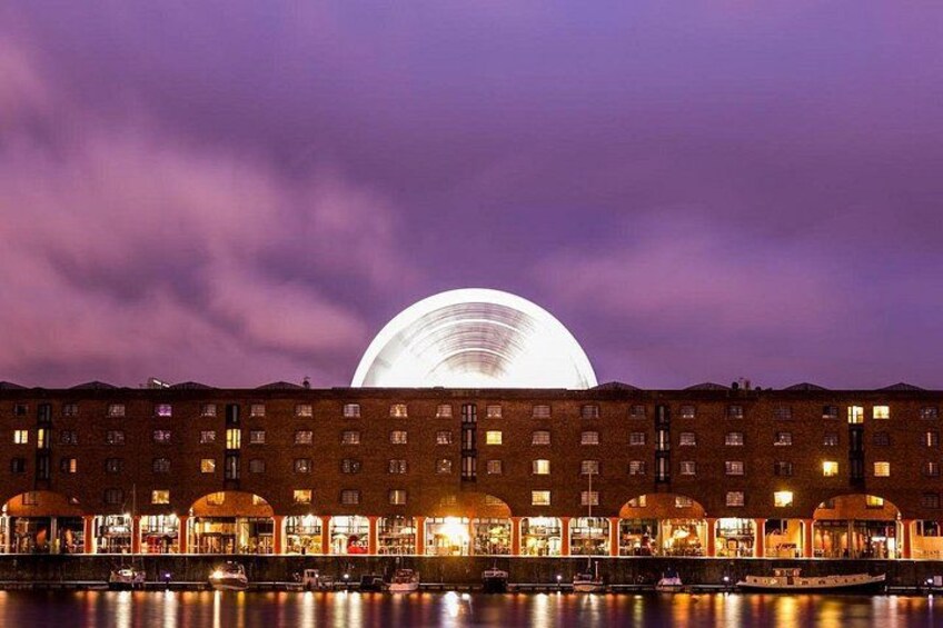 A waterfront building with illuminated arches and a large circular structure in the background. 3