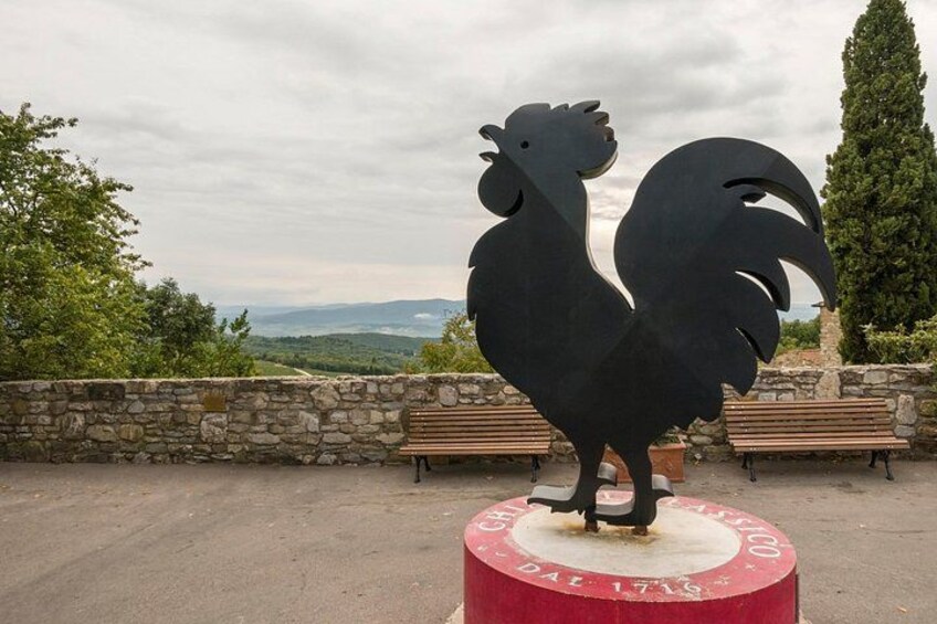 A rooster statue on a red circular base with inscriptions, set against a stone wall with benches, overlooking a scenic landscape. 2