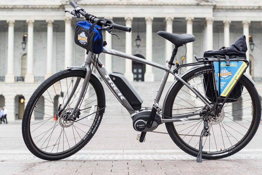 Una bicicleta estática con una bolsa azul y una caja azul etiquetada con '#BIKEROLL' frente a un edificio clásico con columnas. 5