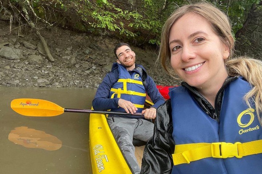 Two people in a yellow kayak on a river, wearing life jackets and a blue vest with a logo. 2