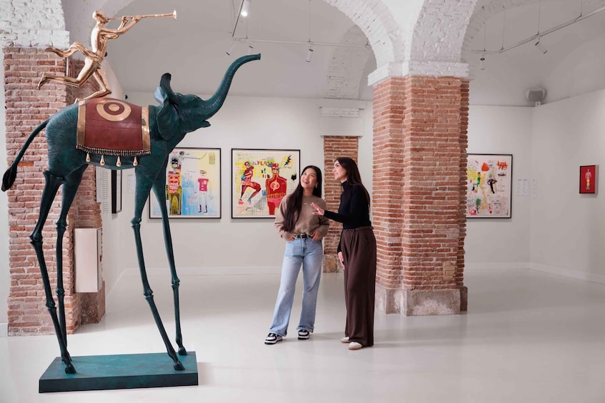 An art gallery with a large sculpture of a figure playing a trumpet on an elephant, and two people examining a painting. 1
