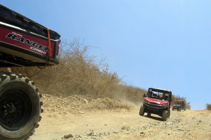 A red off-road vehicle on a dirt road with a large tire, a black vehicle in the background, and dry vegetation. 5