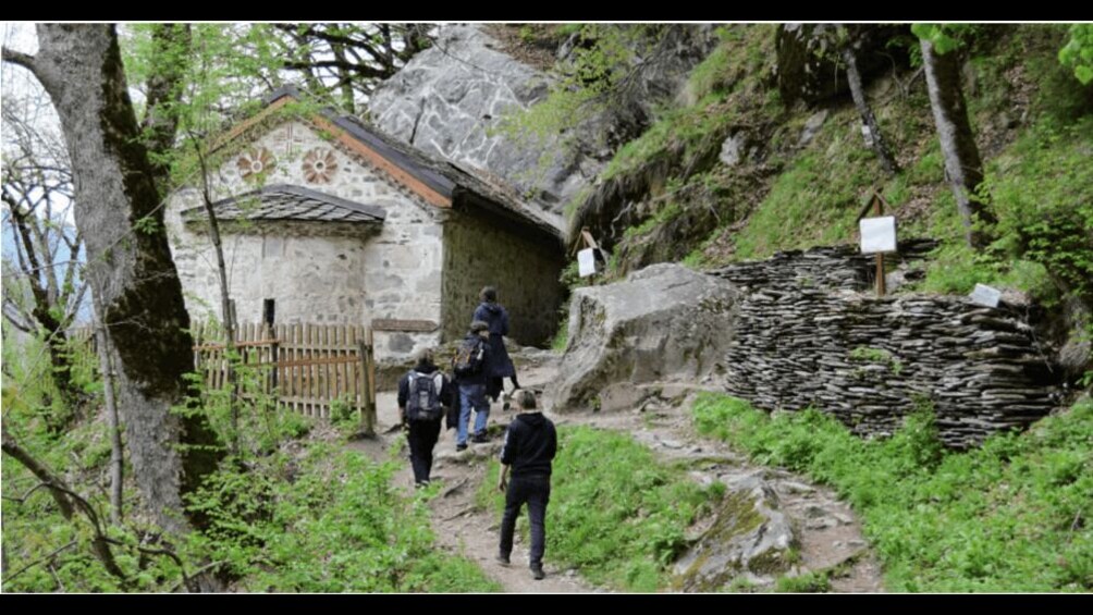 Un edificio de piedra con cerca de madera, excursionistas en un sendero y un muro de piedra. 2