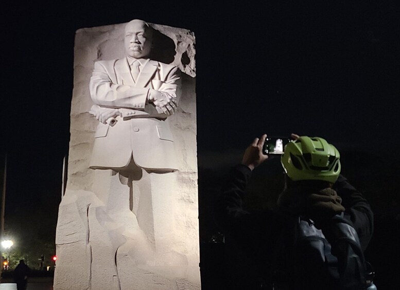 Escultura nocturna de un hombre con traje y los brazos cruzados, siendo fotografiado por una persona que lleva un casco verde. 5