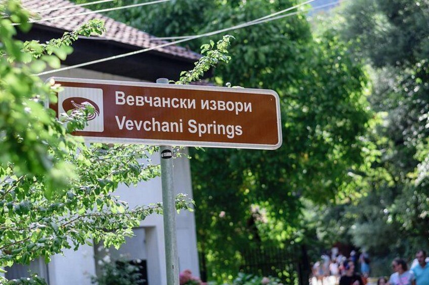 A sign for Vevchanski Izvori, also known as Vechchani Springs, with a backdrop of greenery and a building. 3