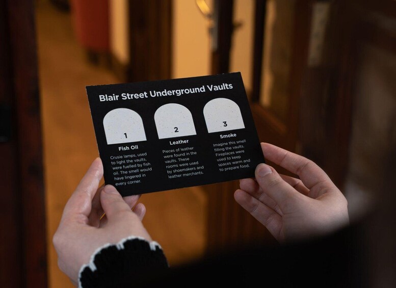 A hand holding a card titled 'Blair Street Underground Vaults' with numbered sections and descriptions. 2