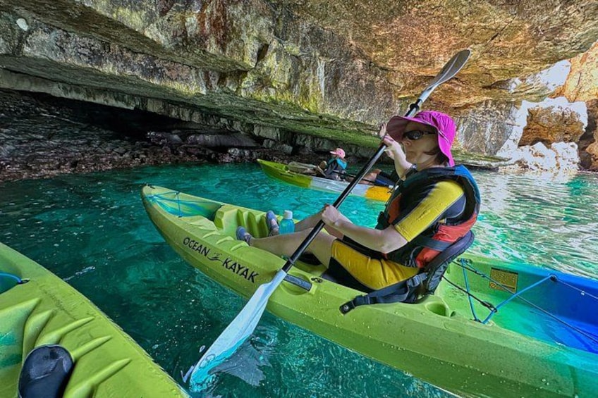 A person kayaking in a green kayak with 'OCEAN KAYAK' written on it, surrounded by clear blue water and rocky cave walls. 4