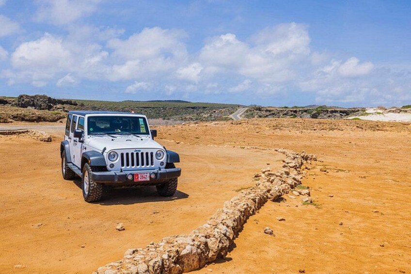 A white off-road vehicle parked on a dirt path with a rocky border, surrounded by a barren landscape with distant hills under a clear sky. 1
