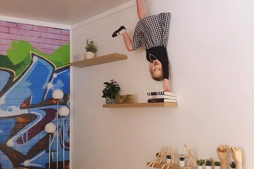 Une personne suspendue la tête en bas dans une pièce ornée d’une fresque colorée, avec des étagères remplies de plantes et de livres. 5