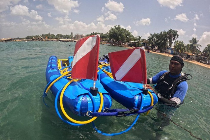 Una persona su una zattera gonfiabile blu e gialla con vele rosse e bianche, che galleggia in acque limpide con una spiaggia e palme sullo sfondo. 3