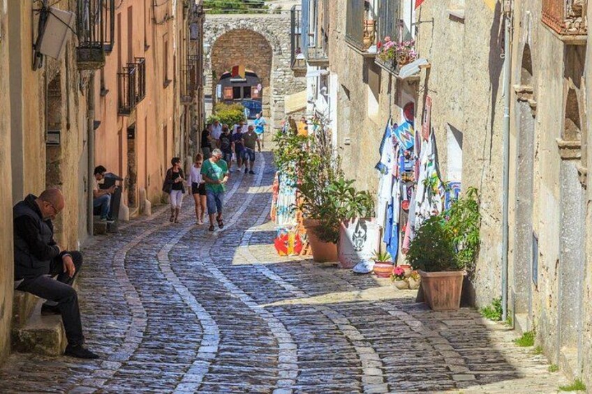 A cobblestone street in a historic town with people walking, a man sitting on a bench, and a shop with colorful items displayed. 3