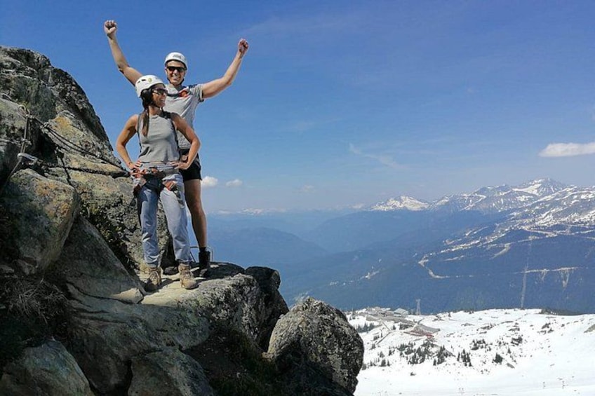 Two people standing on a rocky mountain peak with snow-covered slopes and distant mountains in the background. 2