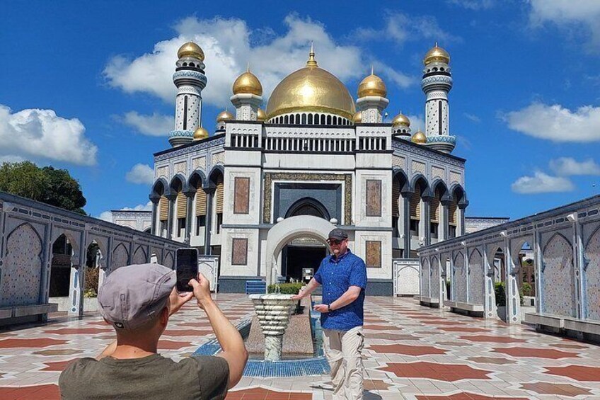 A person taking a photo of a mosque with golden domes and a large central dome, surrounded by a patterned courtyard. 5