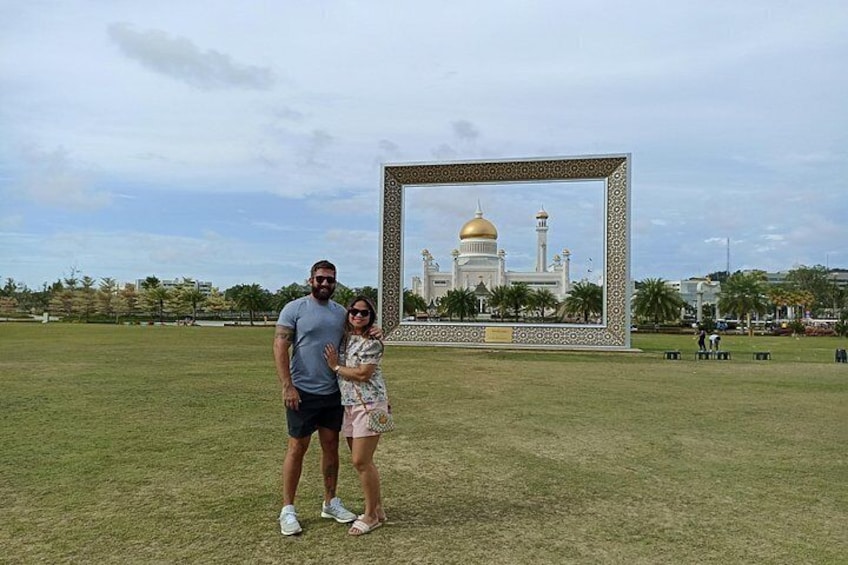 A man and a woman standing in a grassy field with a large framed picture of a white building with domes in the background. 4