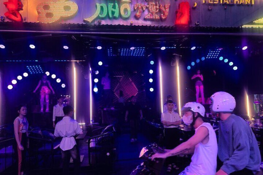 A DJ booth with people, a stage with performers, and a neon sign above. 3
