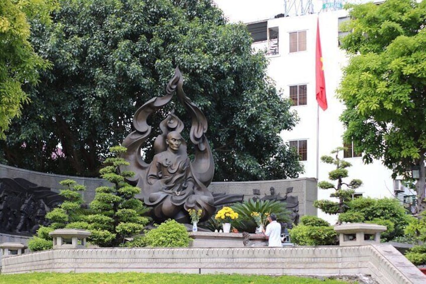 A bronze statue of a seated figure with a raised hand, surrounded by greenery and a stone wall. 4